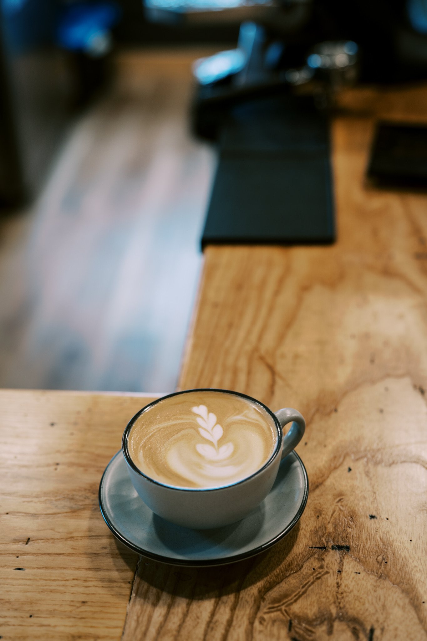 A latte in a cafe cup on a saucer with latte art. The cup is sitting on a wooden countertop.