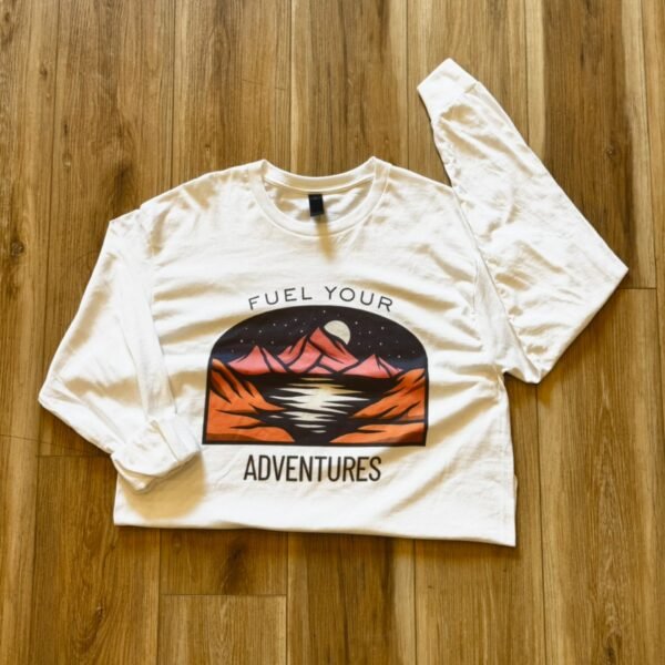 White long sleeve shirt with a beautiful mountain scene with a lake in front of the mountain and the moon partially hidden by the mountain peak. Fuel your adventures is written on the shirt